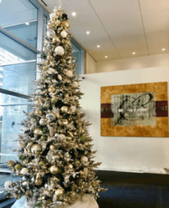 A tall flocked Christmas tree decorated with gold, silver, and white ornaments in a modern office lobby.