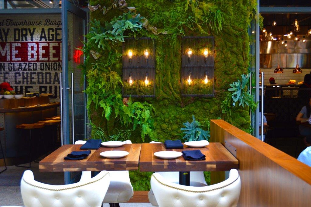 Faux green wall with decorative plants and wall sconces behind a restaurant table setting.
