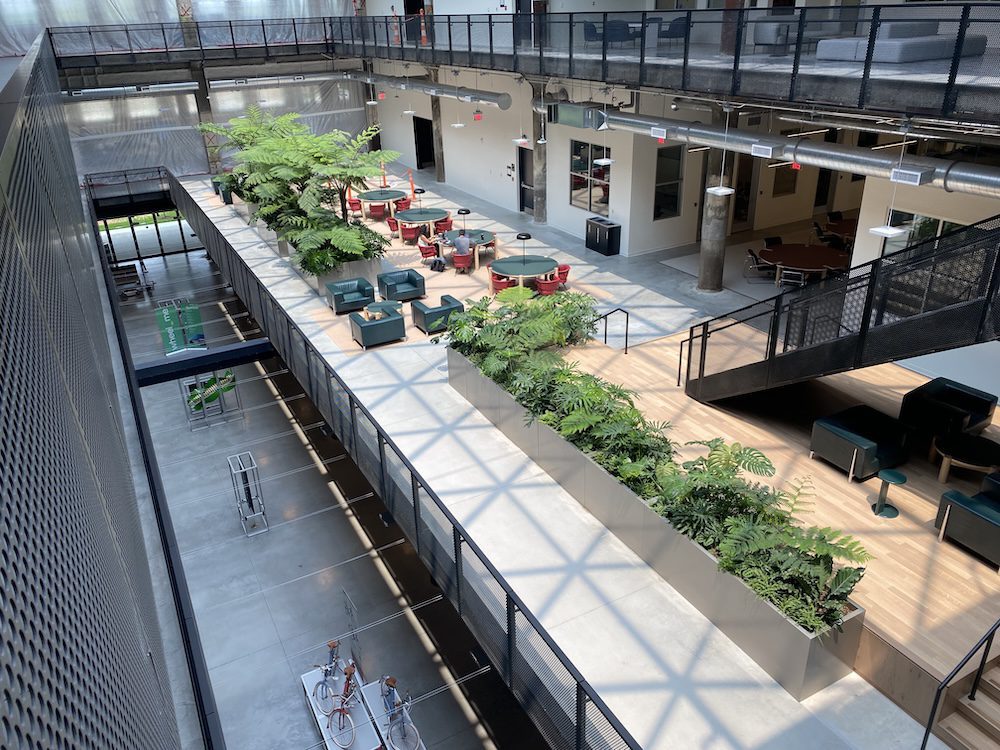 Modern office atrium with lounge seating, plants, and upper walkway overlooking a lower level.